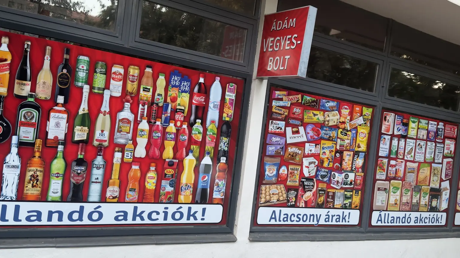 Ádám Vegyesbolt kirakata sűrű termékkollázsokkal, Alacsony árak és Állandó akciók feliratokkal