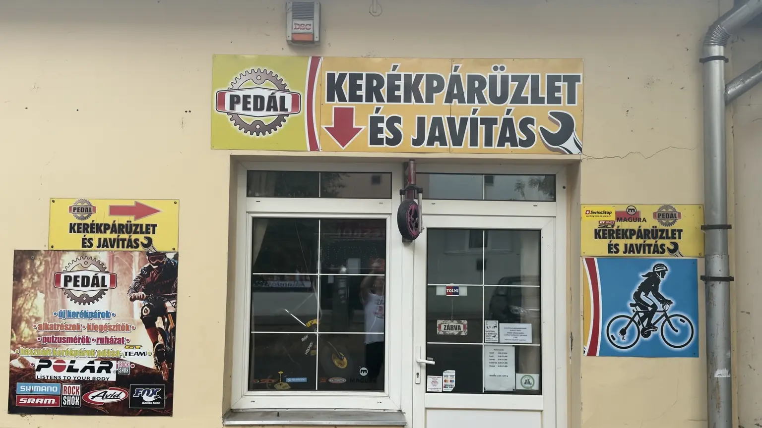 Pedál kerékpárüzlet és javítás homlokzata nagy felirattal, nyilakkal, kerékkel, kerékpáros képekkel és alkatrészmárkákkal