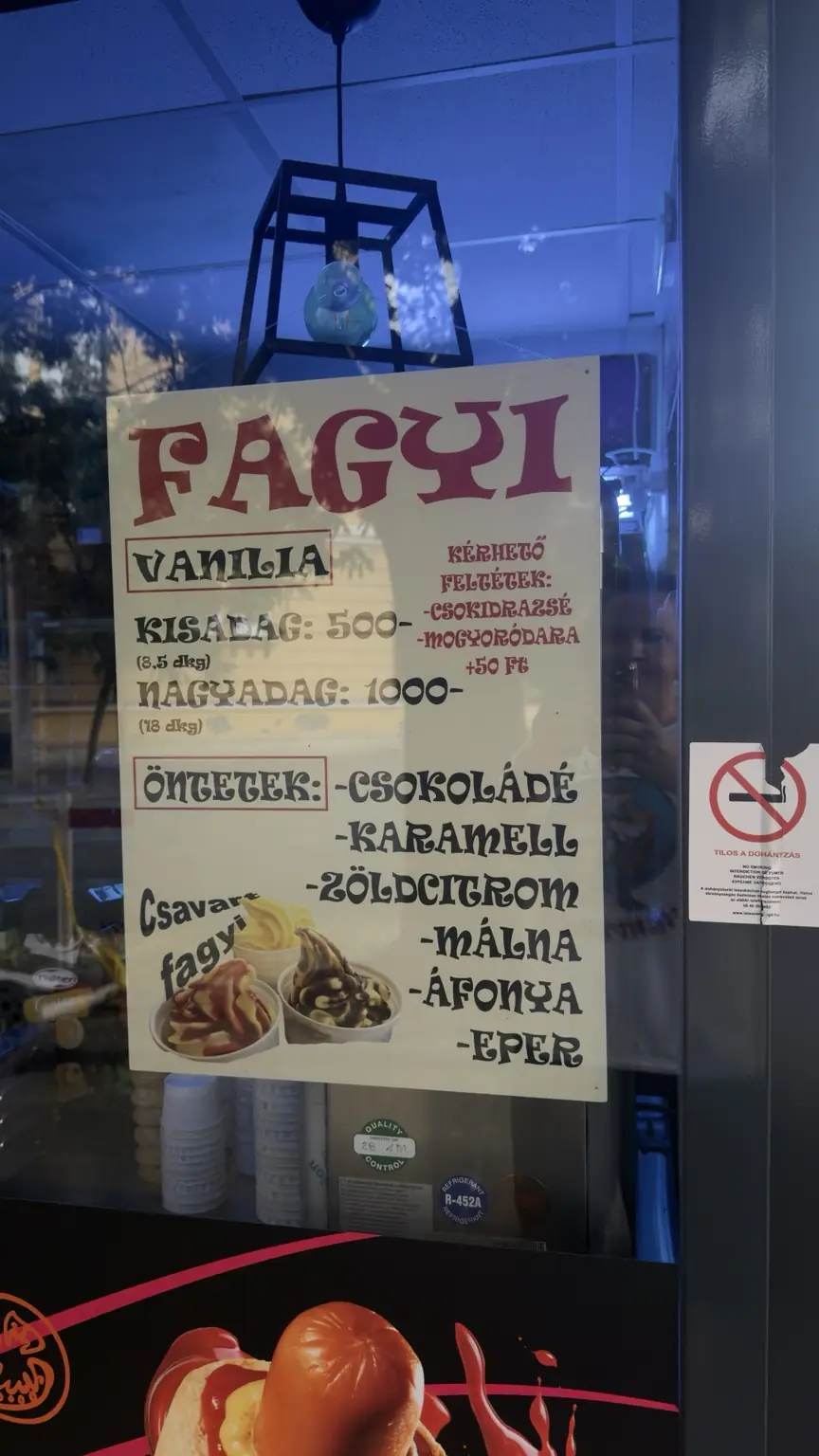 Kézzel írt fagylaltárlista két adagmérettel, ízválasztékkal és feltétárral egy üzlet kirakatában