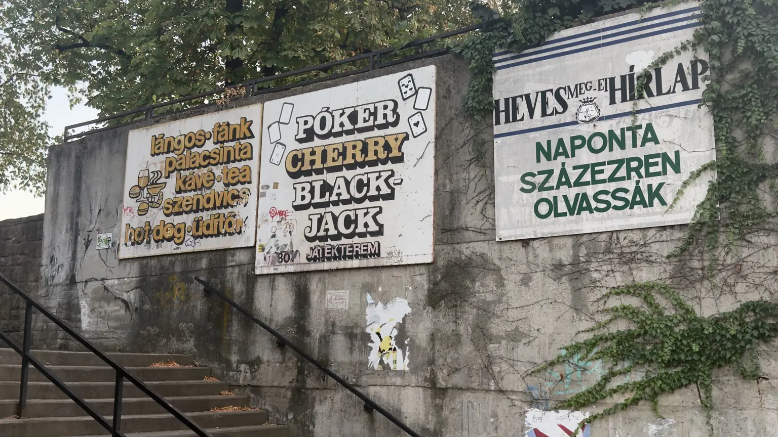 Három egymás melletti, időjárás által megviselt utcai reklámtábla vendéglátóhelyi, játéktermi és újsághirdetéssel