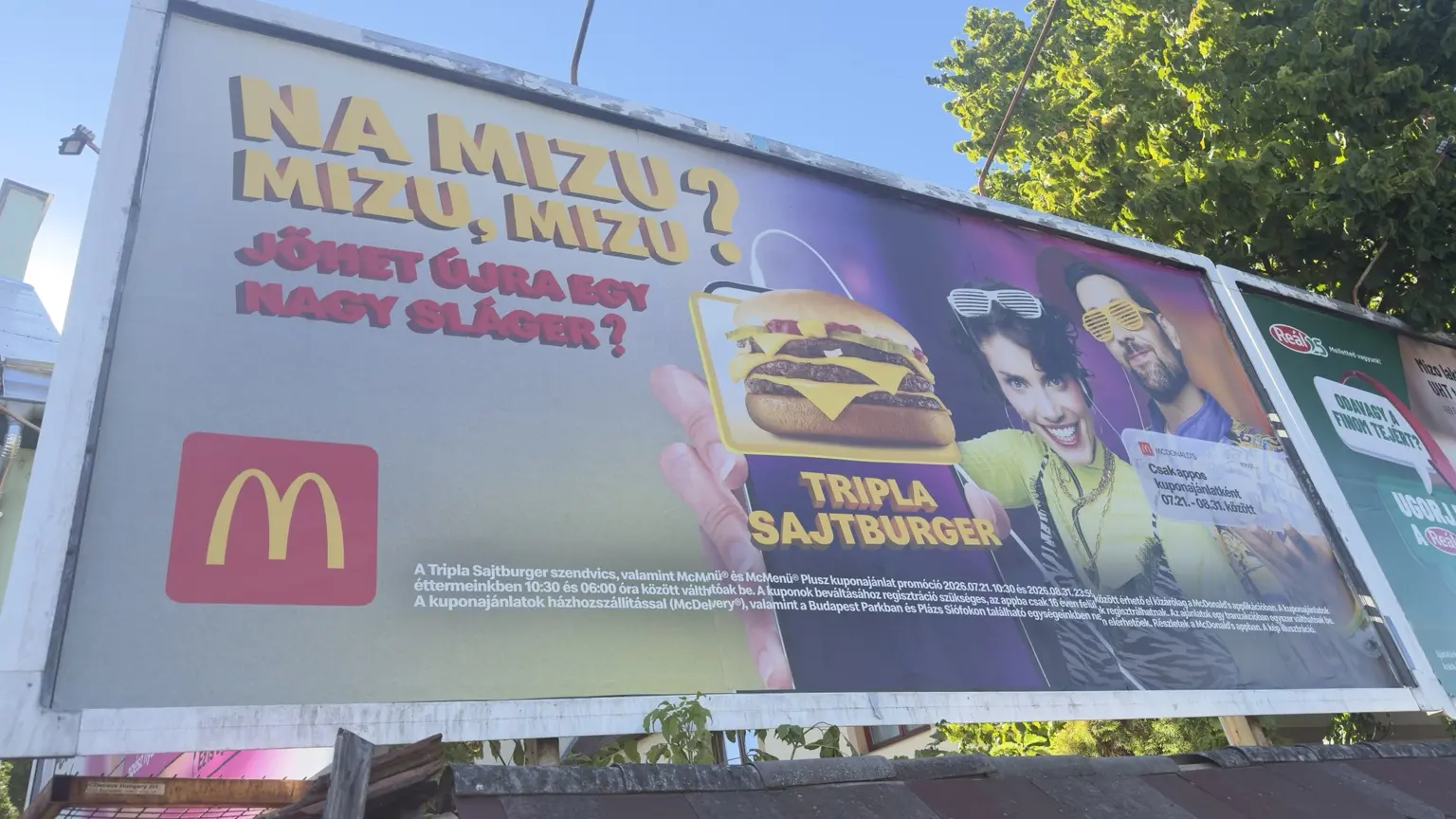 McDonald’s óriásplakát Na mizu szöveggel, két szereplővel, Tripla Sajtburgerrel és appos kuponjelzéssel