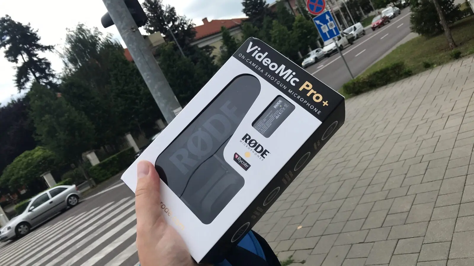Kézben tartott RØDE VideoMic Pro+ mikrofoncsomagolás városi járda és út előtt